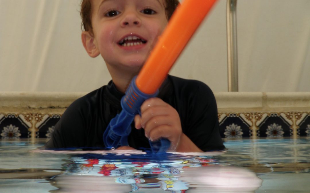 A young boy smiling in a pool holding an orange water toy.