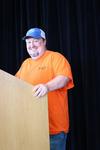 Man in orange shirt speaking at a podium on stage.