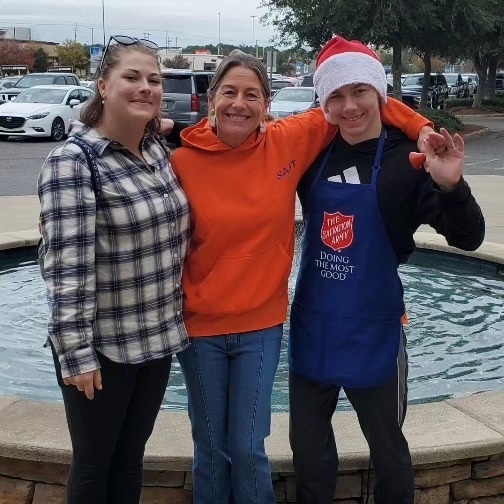 Three people smiling by a waterfront, one wearing a Santa hat and Salvation Army apron.