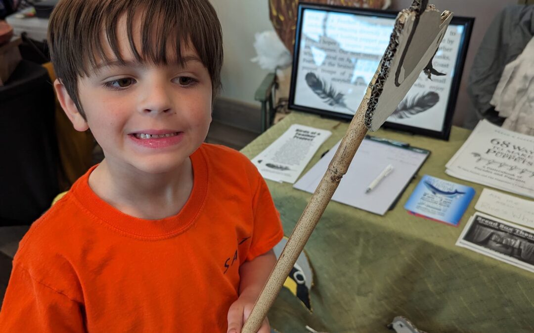 A smiling boy in an orange shirt holding a wooden stick indoors.